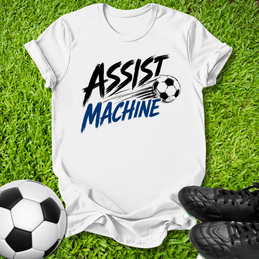A white short-sleeve graphic T-shirt laid flat on green grass, featuring the words 'ASSIST MACHINE' in black and blue with a soccer ball illustration. A black-and-white soccer ball and black cleats are placed around it as props.