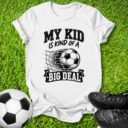 A white short-sleeve graphic T-shirt laid flat on green grass, with a soccer ball and black cleats nearby. The shirt has a black printed slogan reading 'MY KID IS KIND OF A BIG DEAL' around a soccer ball and net graphic.