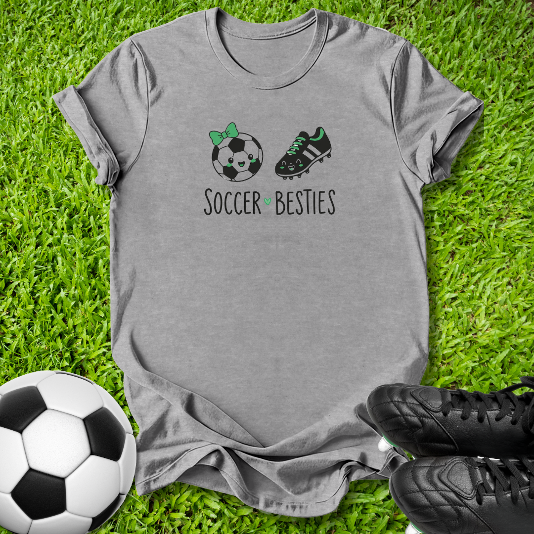 A heather gray short-sleeve t-shirt laid flat on green grass, featuring a graphic of a soccer ball with a green bow, a black soccer cleat, and the text 'SOCCER BESTIES'. A black-and-white soccer ball is on the bottom left, and black cleats are on the bottom right.