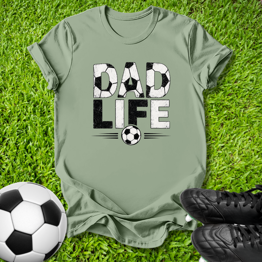 An olive green t-shirt with a 'DAD LIFE' soccer graphic, laid flat on green grass with a soccer ball and black cleats as props.