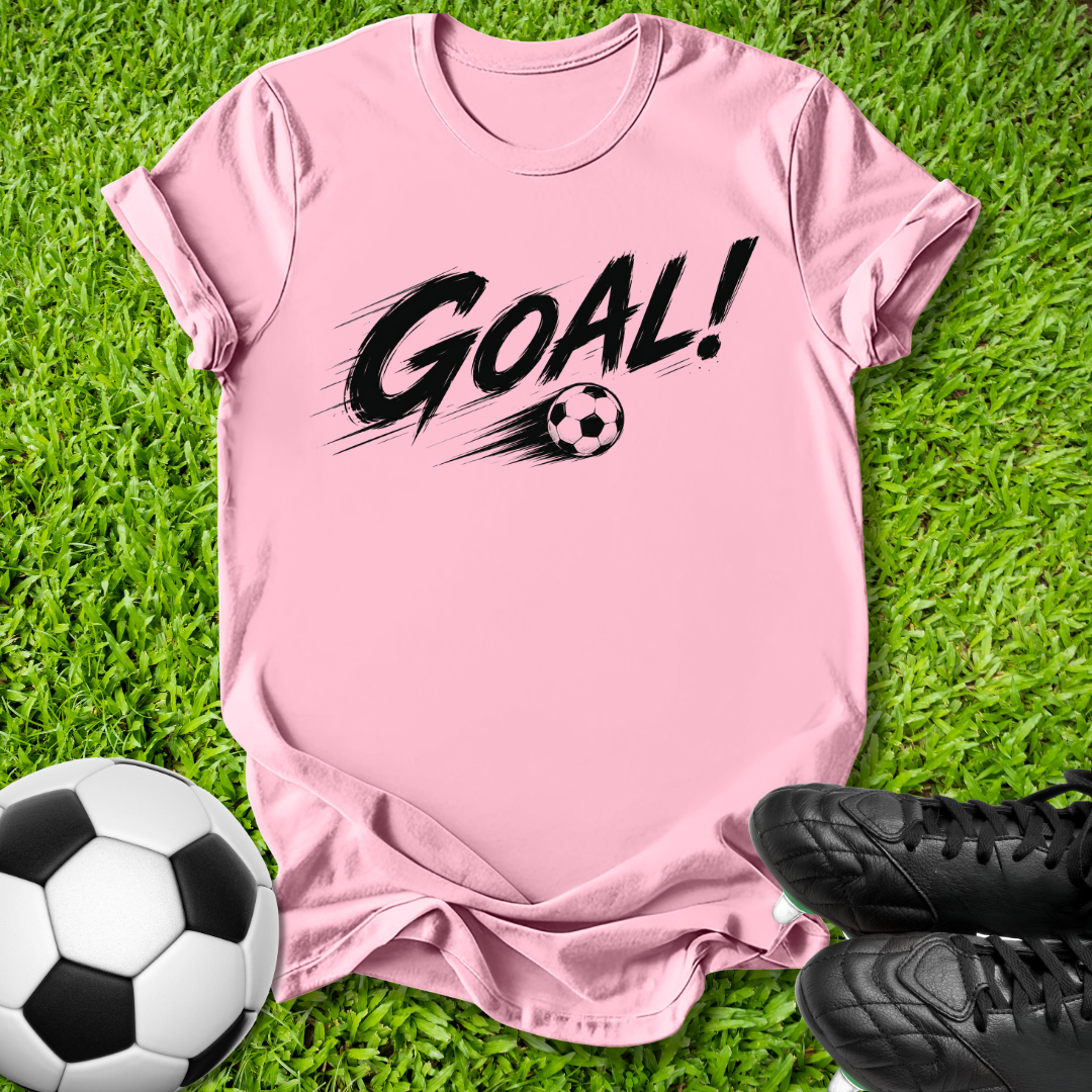 A pink short-sleeve t-shirt with a black 'GOAL!' graphic and soccer ball print, laid flat on green grass with a soccer ball and black cleats as props.