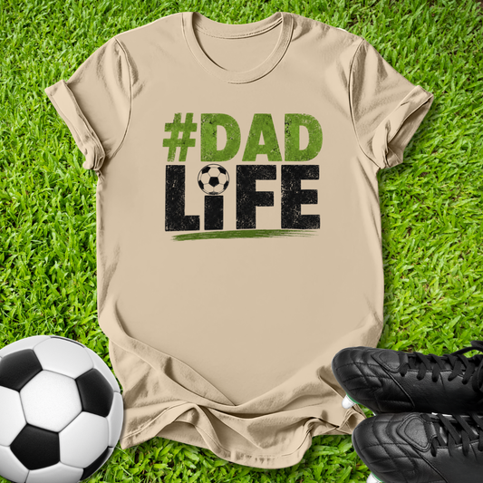 A beige graphic t-shirt laid flat on green grass, featuring the text '#DAD LIFE' in distressed green and black lettering with a soccer ball motif. A soccer ball and black cleats are placed around it as props.