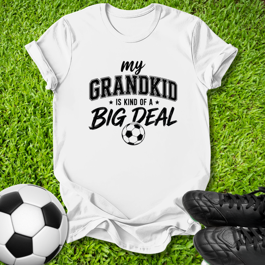 A white short-sleeve graphic T-shirt laid flat on green grass, with a soccer ball and black cleats nearby. The shirt has black text reading 'my GRANDKID IS KIND OF A BIG DEAL' with a small soccer ball graphic.