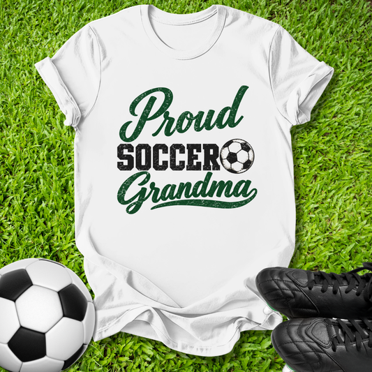 A white short-sleeve T-shirt laid flat on green grass, printed with the words 'Proud Soccer Grandma' in dark green and black, with a soccer ball graphic. A soccer ball and black cleats are placed around it as props.