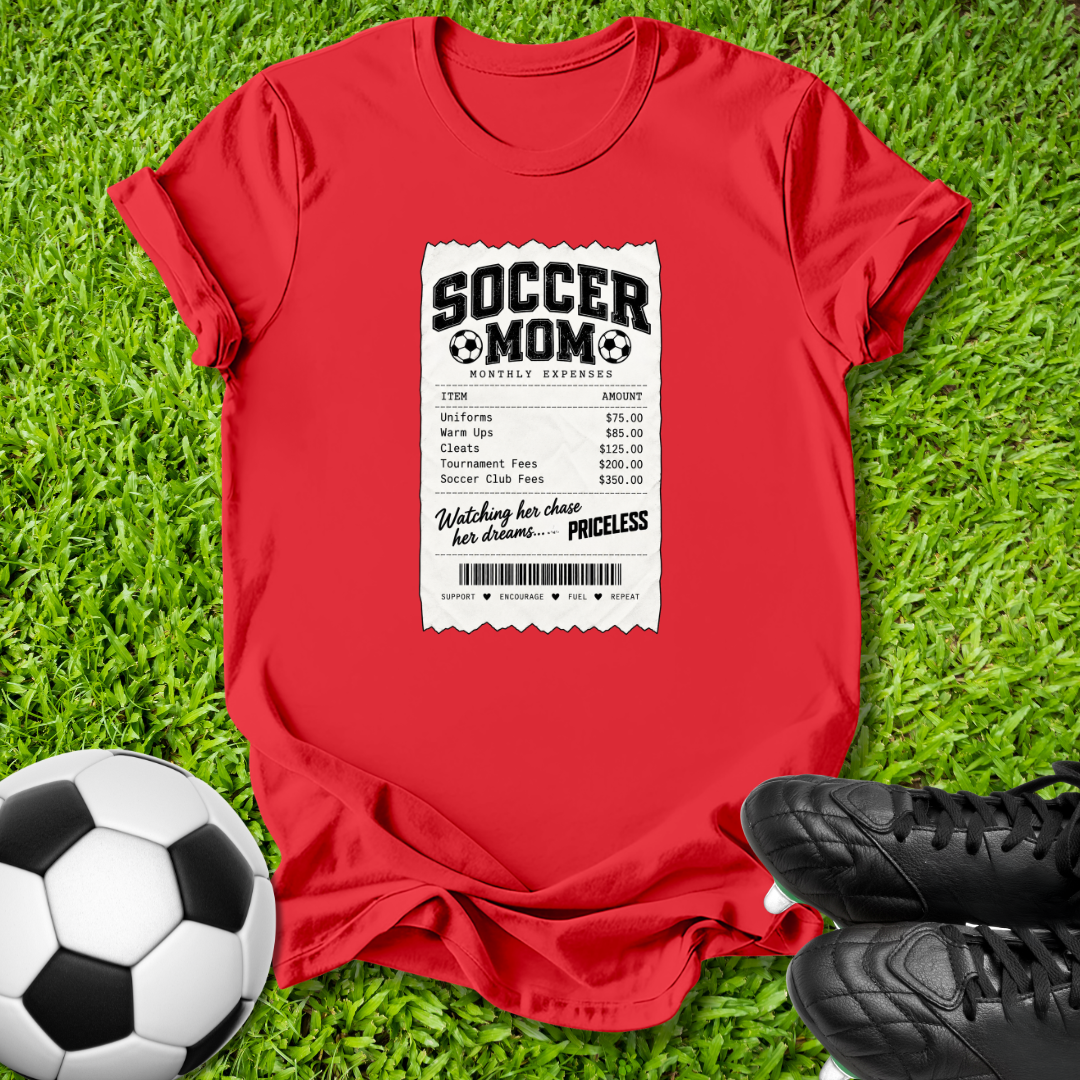 A red short-sleeve graphic T-shirt laid flat on green grass, featuring a white ticket-style print that reads 'SOCCER MOM' with a receipt layout. A soccer ball is at the lower left and black cleats are at the lower right.