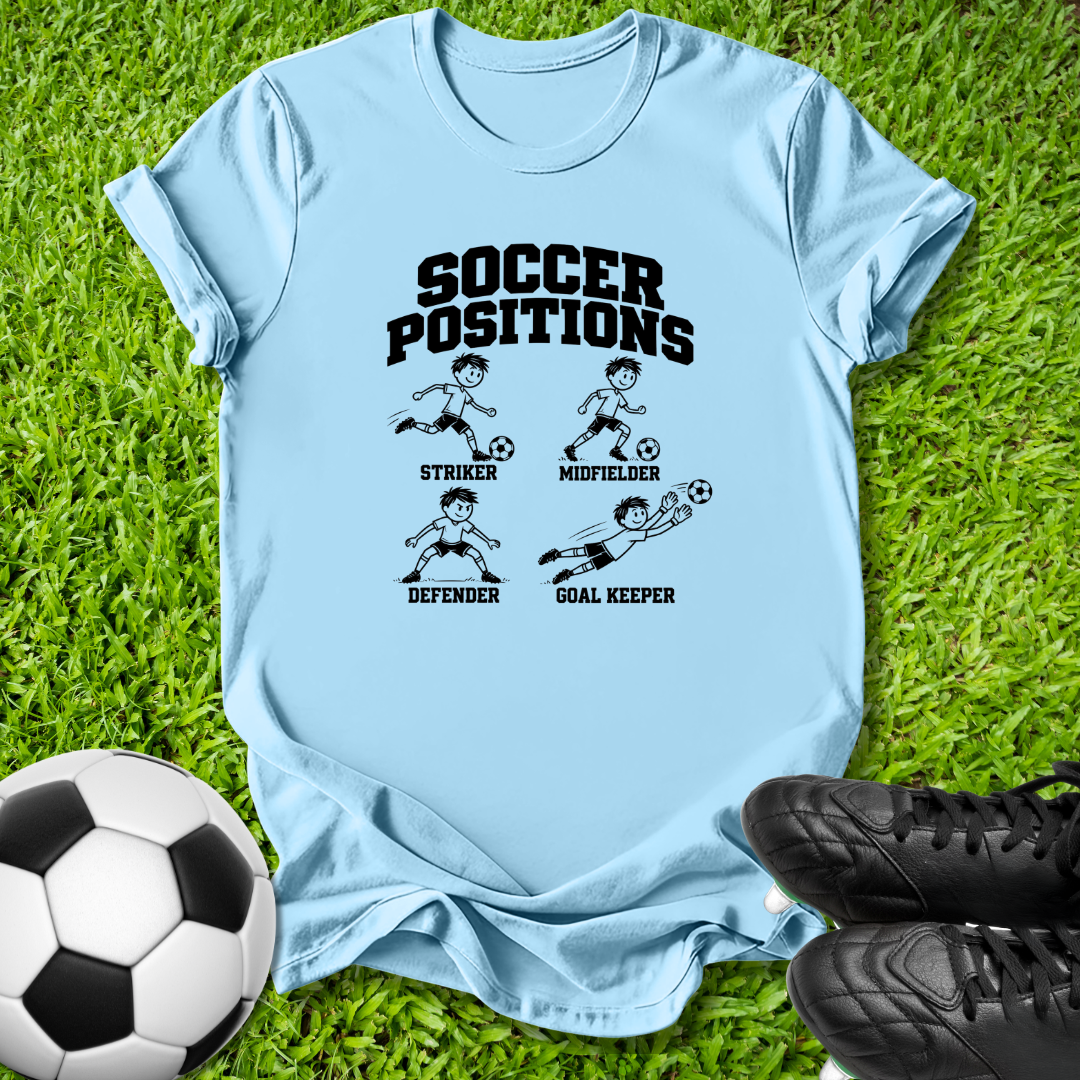 A light blue graphic t-shirt laid flat on green grass, featuring a black 'SOCCER POSITIONS' print with illustrations of a striker, midfielder, defender, and goal keeper. A soccer ball is on the bottom left and black cleats are on the bottom right.