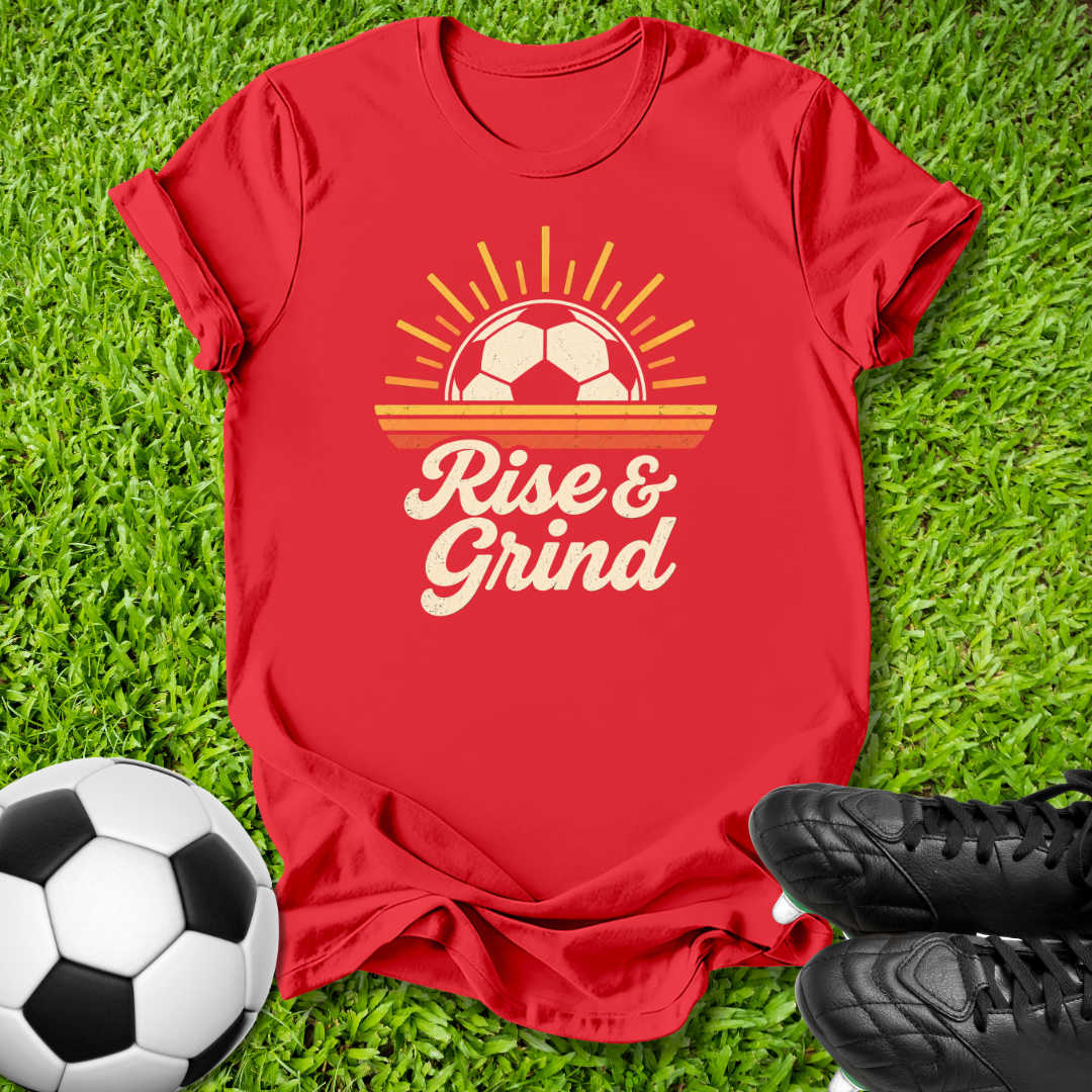 A red short-sleeve graphic T-shirt laid flat on green grass, featuring a retro soccer-themed design with a soccer ball, sun rays, and the words 'Rise & Grind.' A soccer ball and black cleats are placed around it as styling props.