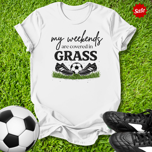 White t-shirt with 'my weekends are covered in grass' text and soccer ball graphics on grass with soccer shoes and ball.