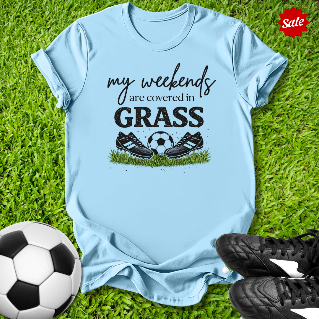 Light blue t-shirt with 'my weekends are covered in grass' text and soccer ball graphic on grass with soccer shoes and ball.