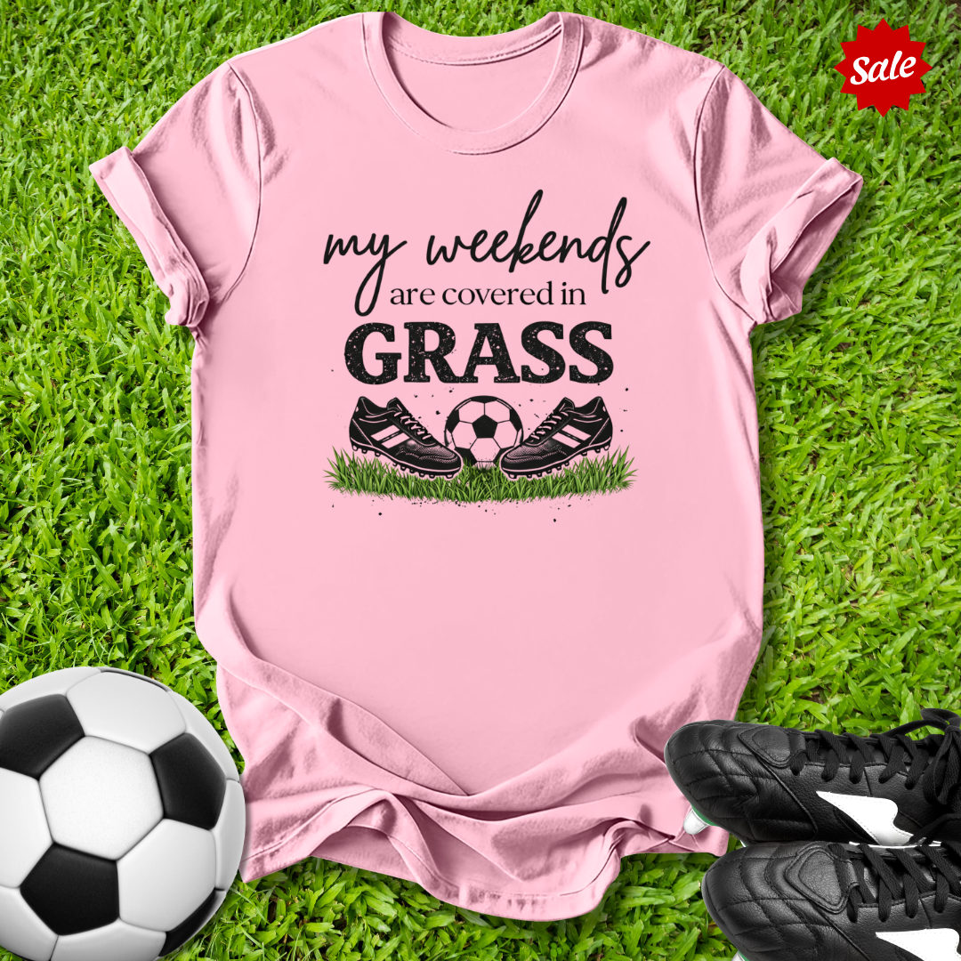 Pink t-shirt with 'my weekends are covered in grass' text and soccer ball graphics on grass with soccer shoes and ball.