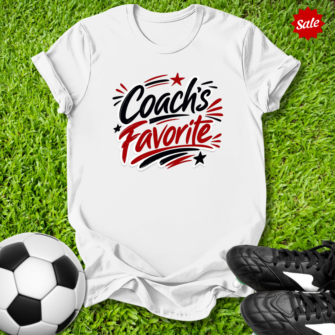 White t-shirt with 'Coach's Favorite' text on grass with soccer ball and shoes