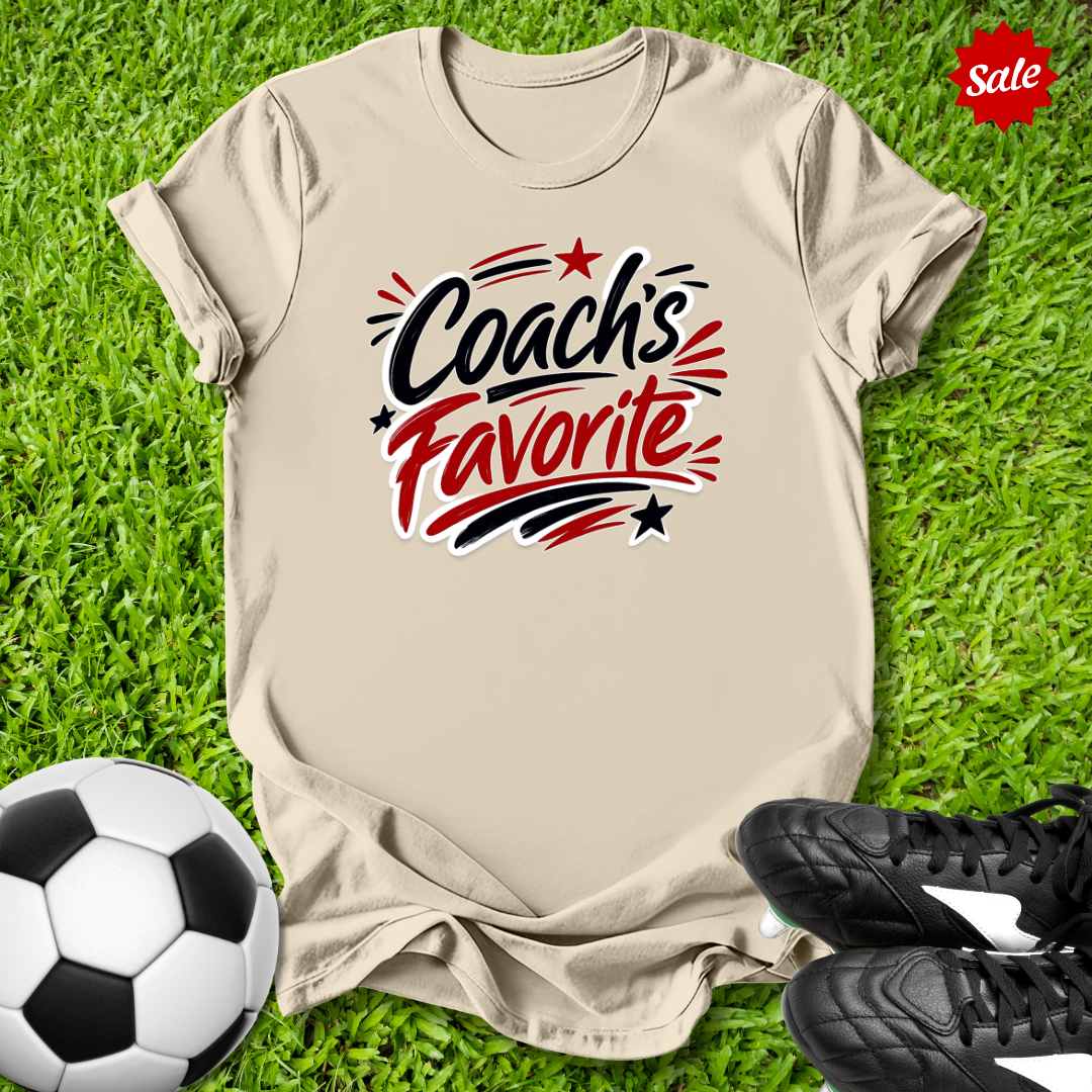 Beige t-shirt with 'Coach's Favorite' text on grass with soccer ball and shoes