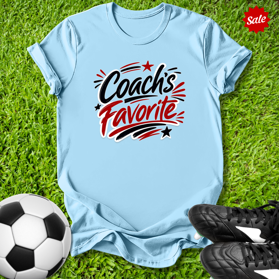 Light blue t-shirt with 'Coach's Favorite' text on grass with soccer ball and shoes