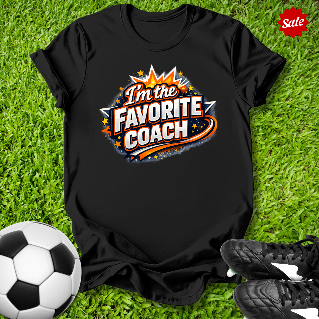 Black t-shirt with 'I'm the Favorite Coach' design on grass with soccer ball and shoes.