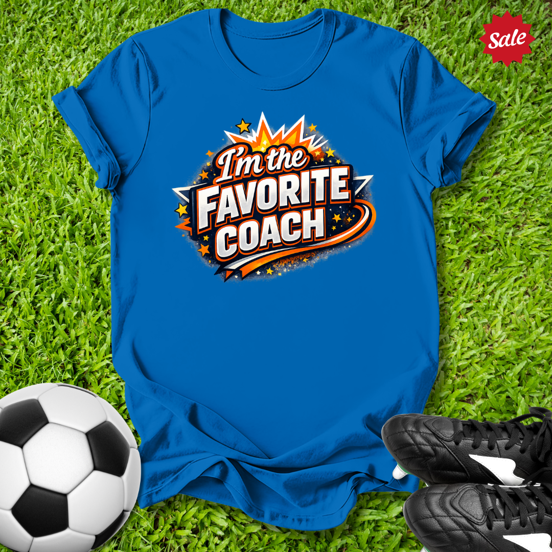 Blue t-shirt with 'I'm the Favorite Coach' design on grass with soccer ball and shoes.