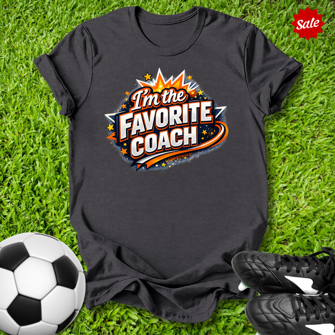 Gray t-shirt with 'I'm the Favorite Coach' design on grass with soccer ball and shoes.