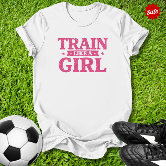 White t-shirt with 'Train Like a Girl' text on grass with soccer ball and shoes