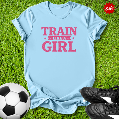 Light blue t-shirt with 'Train Like a Girl' text on grass with soccer ball and shoes.