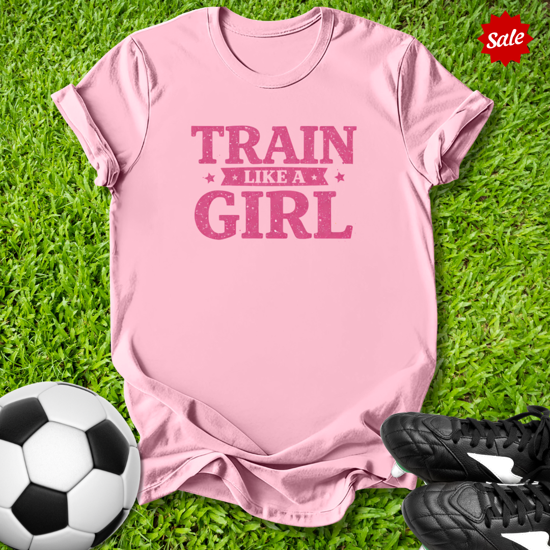 Pink t-shirt with 'Train Like a Girl' text on grass with soccer ball and shoes