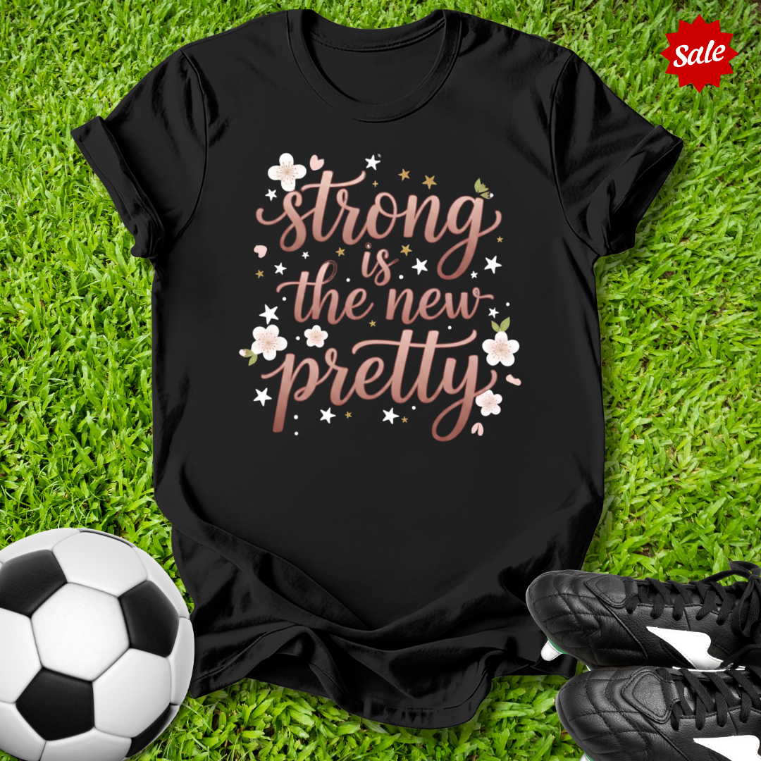 Black t-shirt with 'strong is the new pretty' text on grass with soccer ball and shoes