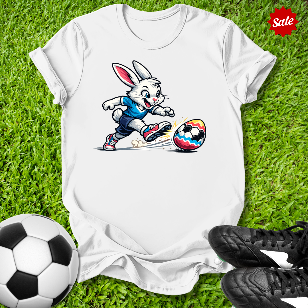 White t-shirt with  the  Easter bunny kicking a soccer ball on grass, surrounded by soccer balls and shoes.