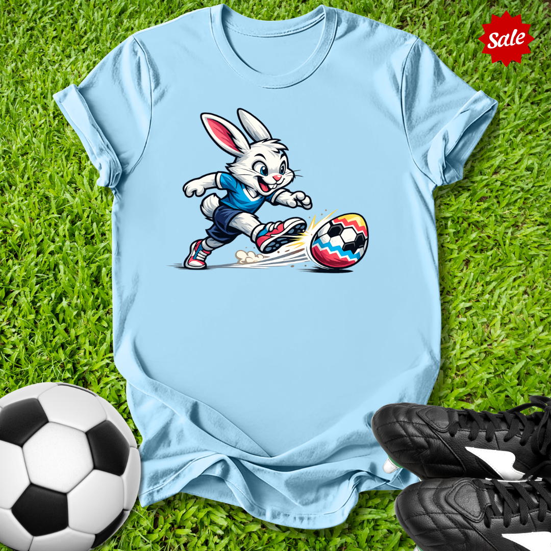 Light blue t-shirt with  the  Easter bunny playing soccer on grass with soccer balls and shoes.
