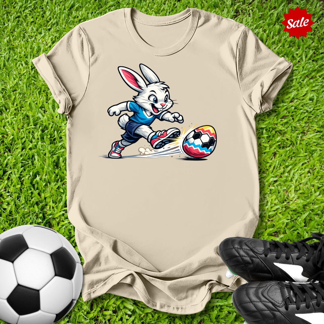 Beige t-shirt with  the  Easter bunny playing soccer on grass with soccer balls and shoes.