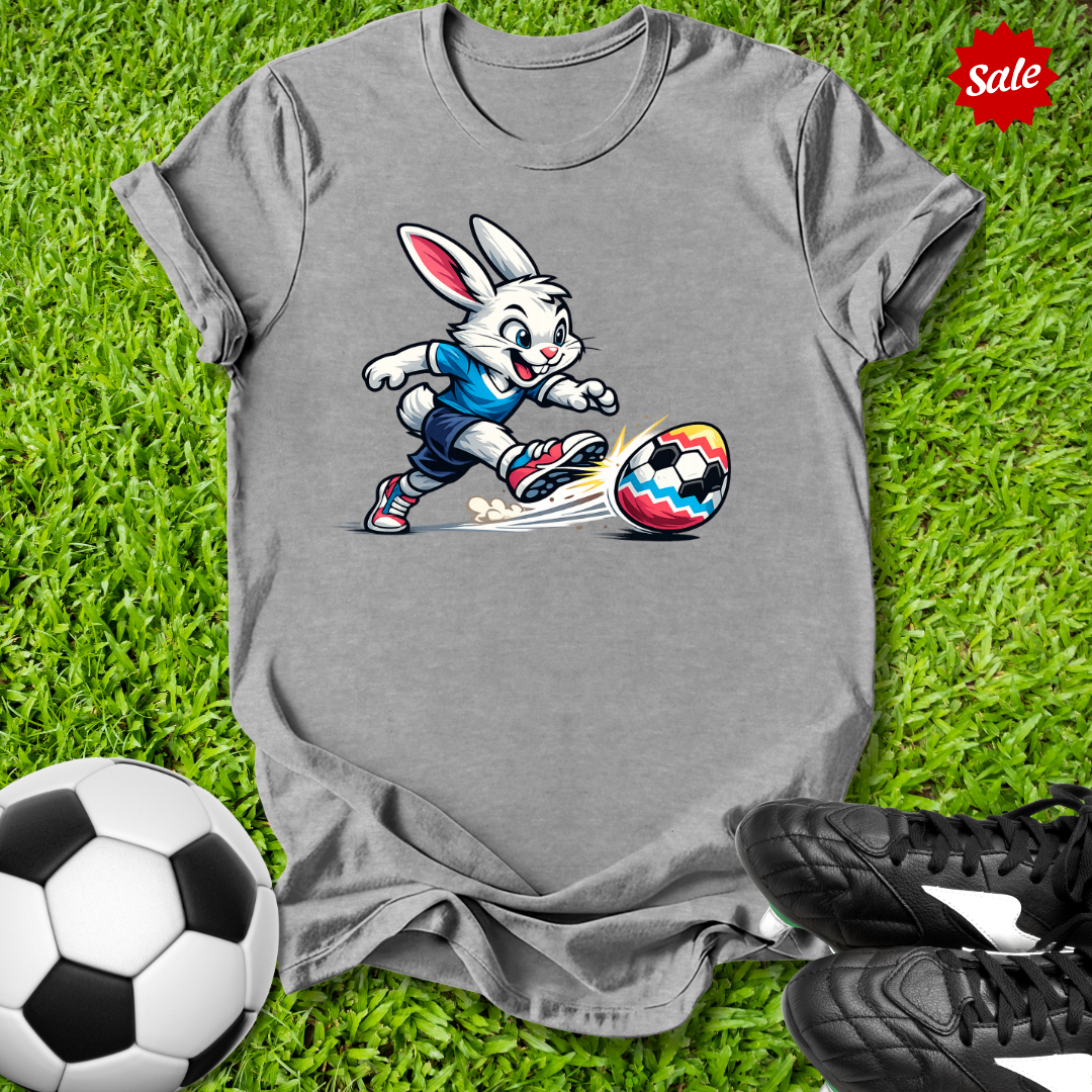 Gray t-shirt with the  Easter bunny kicking a soccer ball on grass, with a soccer ball and shoes in the foreground.
