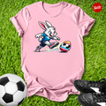 Pink t-shirt with the  Easter bunny kicking a soccer ball on grass, surrounded by soccer balls and shoes.