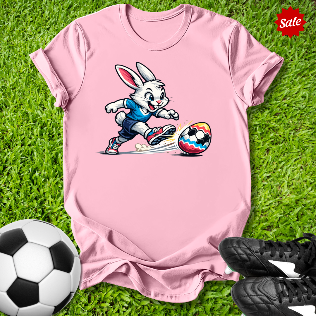 Pink t-shirt with the  Easter bunny kicking a soccer ball on grass, surrounded by soccer balls and shoes.