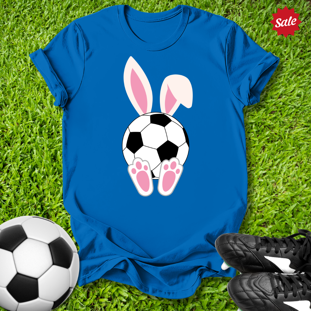 Blue t-shirt with a soccer ball and  Easter bunny ears design on grass, with a soccer ball and shoes around.