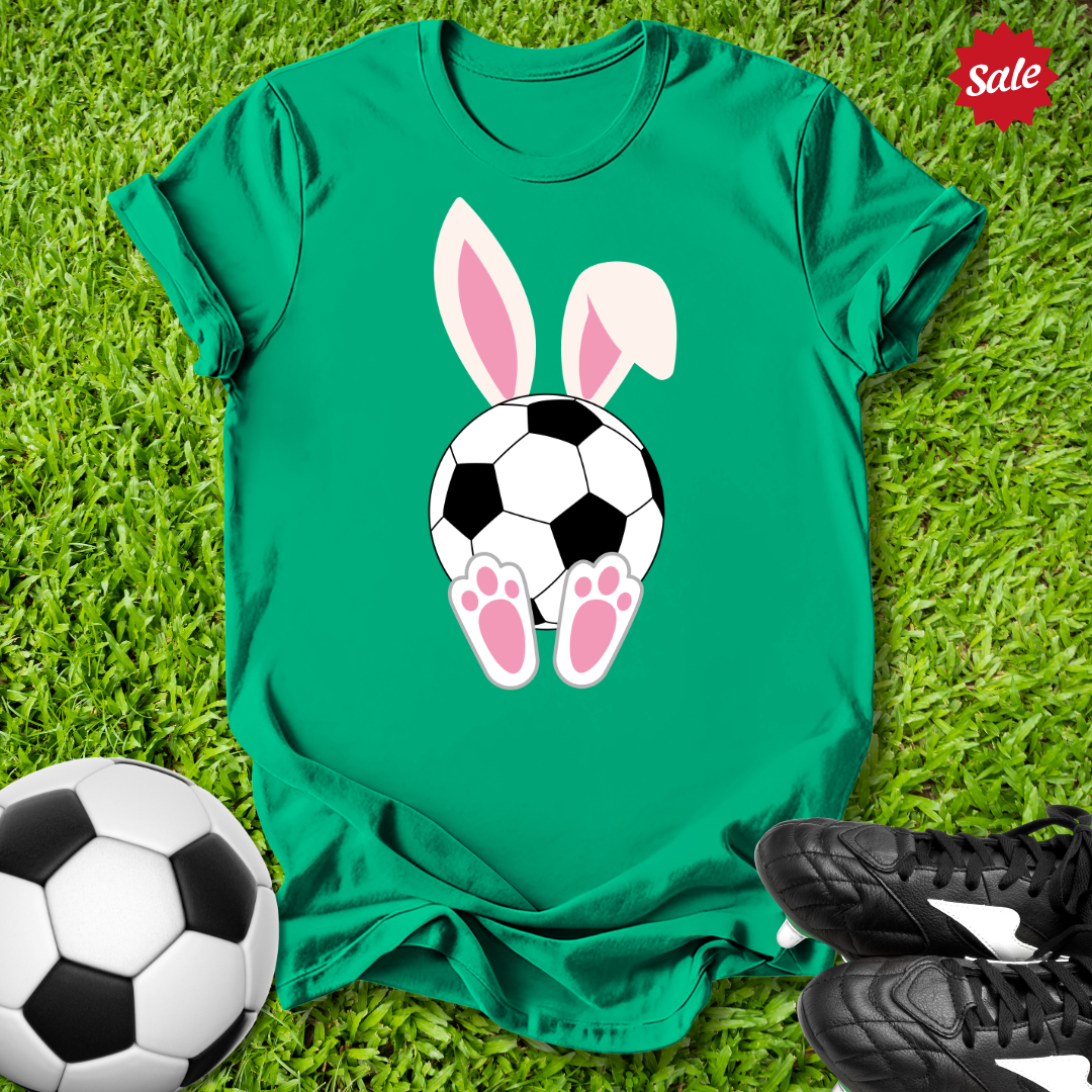 Green t-shirt with Easter bunny ears and soccer ball design on grass, with soccer ball and shoes around.
