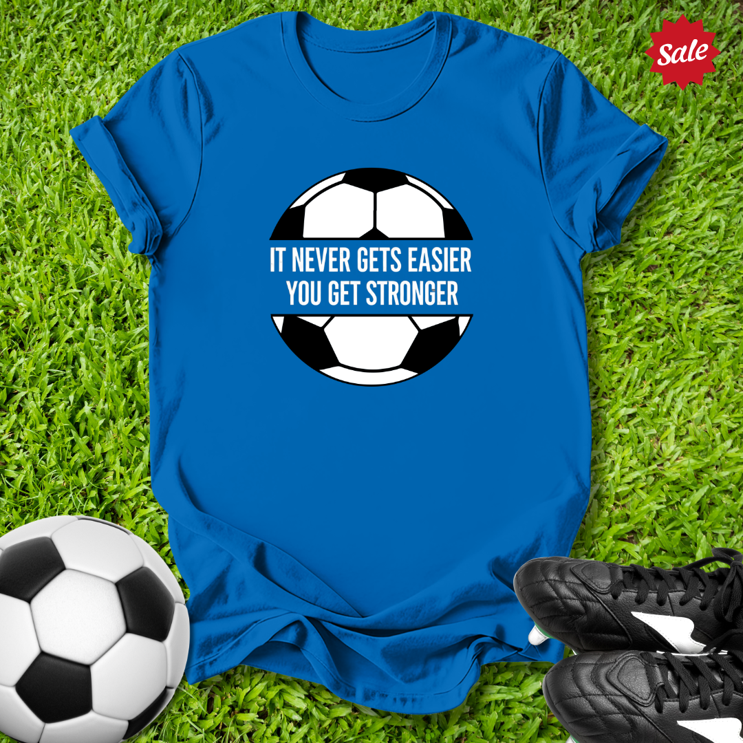 Blue t-shirt with soccer ball graphic and motivational quote on grass with soccer ball and shoes.