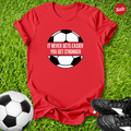 Red t-shirt with soccer ball graphic and motivational quote on grass with soccer ball and shoes.