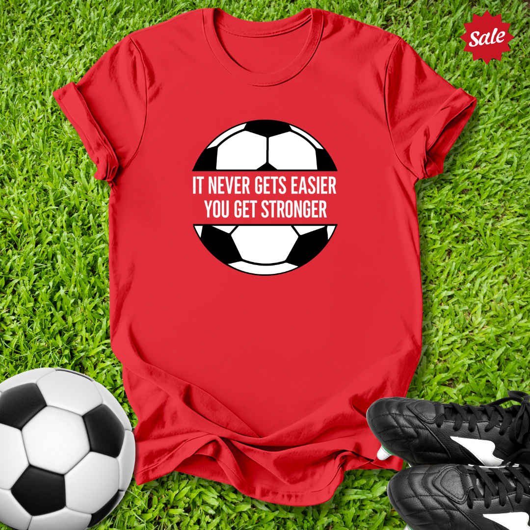Red t-shirt with soccer ball graphic and motivational quote on grass with soccer ball and shoes.
