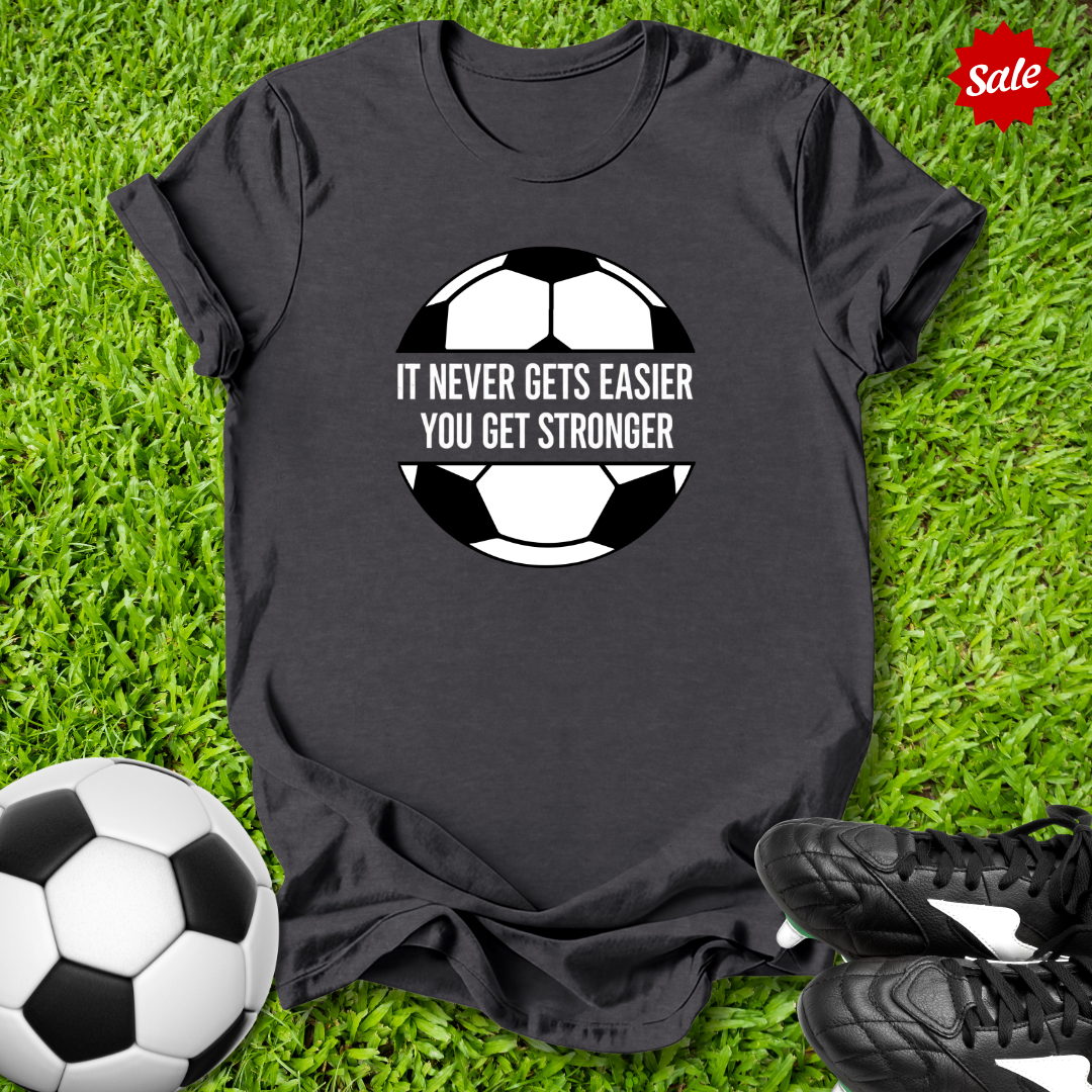 Black t-shirt with soccer ball graphic and motivational quote on grass with soccer ball and shoes.