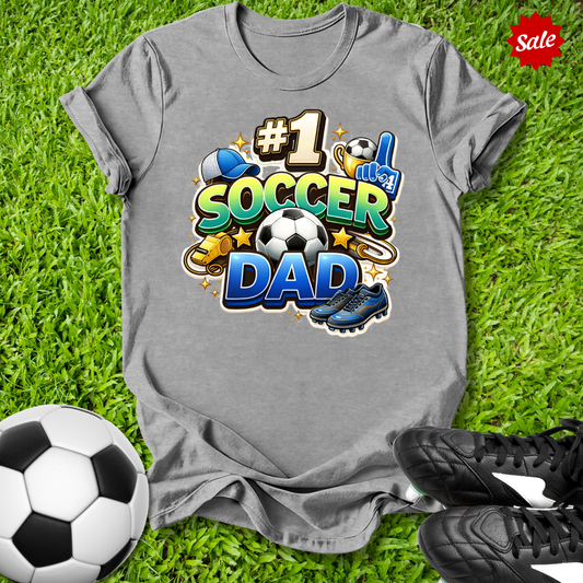 Gray t-shirt with '#1 Soccer Dad' design on grass with soccer ball and cleats.