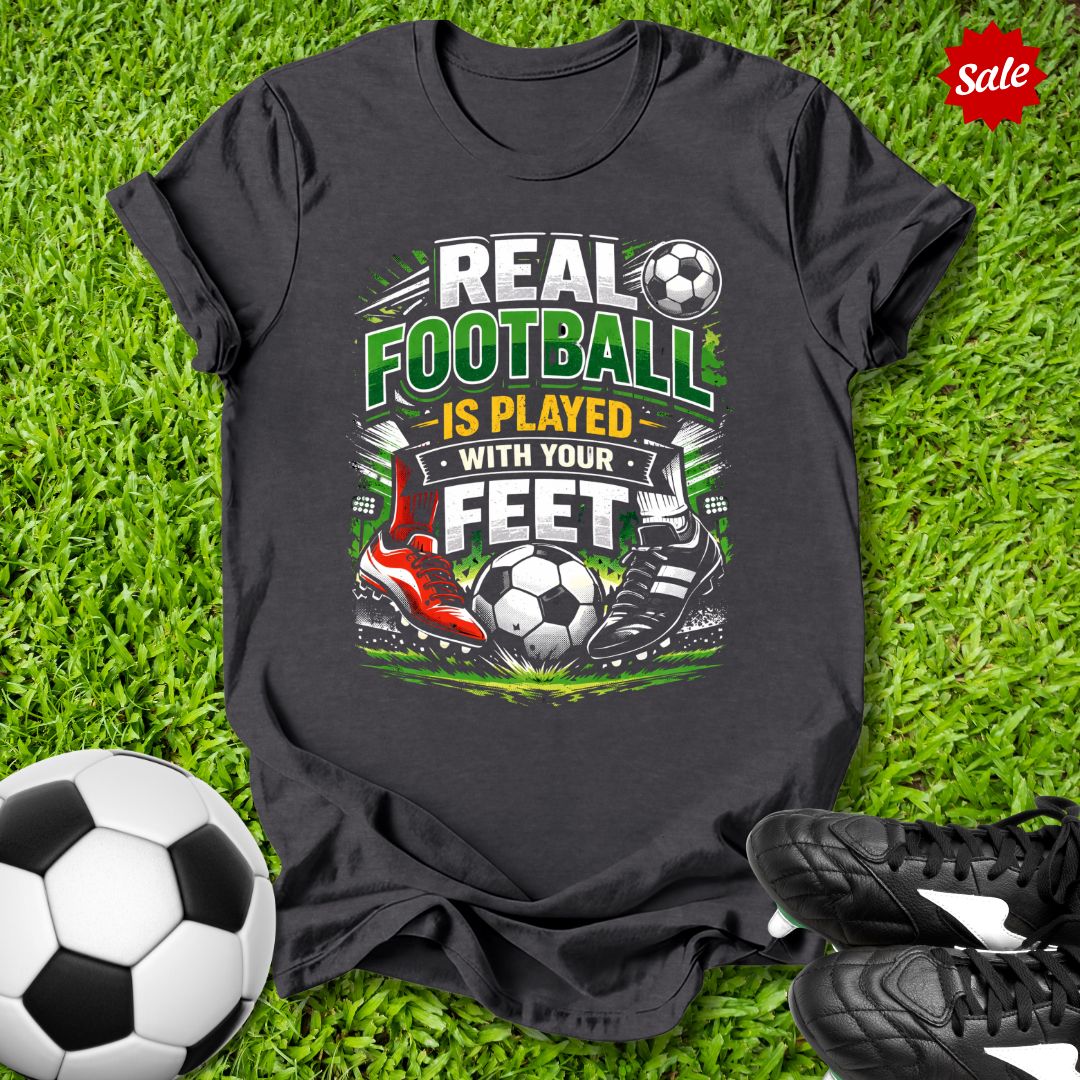 Gray t-shirt with soccer-themed design on grass with soccer ball and shoes