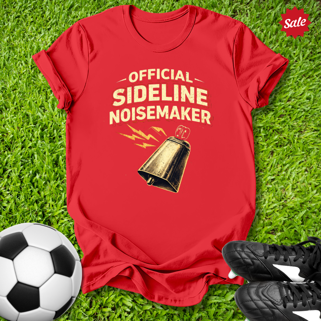 Red t-shirt with 'Official Sideline Noisemaker' design on grass with soccer ball and shoes.