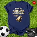 Navy blue t-shirt with 'Official Sideline Noisemaker' design on grass with soccer ball and shoes.
