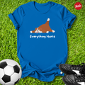 Blue t-shirt with a dog graphic and 'Everything Hurts' text on grass with soccer ball and shoes.