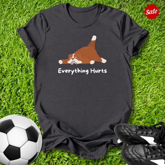 Black t-shirt with a dog graphic and 'Everything Hurts' text on grass with soccer ball and shoes.