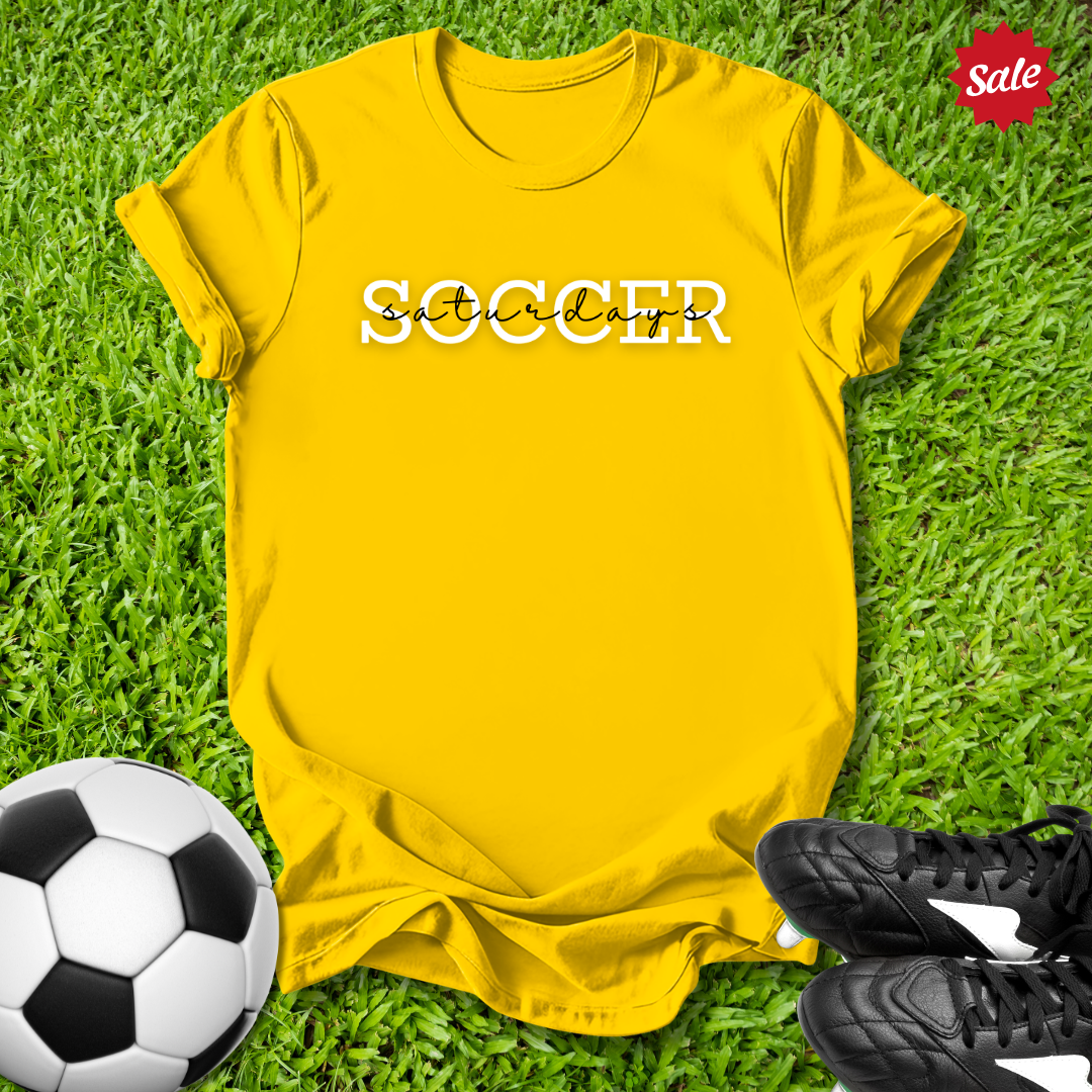 Yellow t-shirt with 'Saturdays' text on grass with soccer ball and shoes