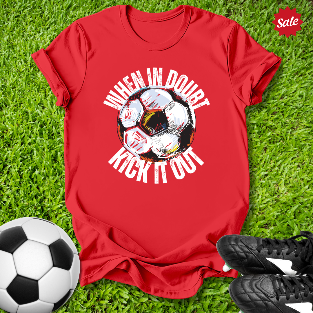 Red t-shirt with  when in doubt kick it out soccer ball graphic and text on grass with soccer ball and shoes