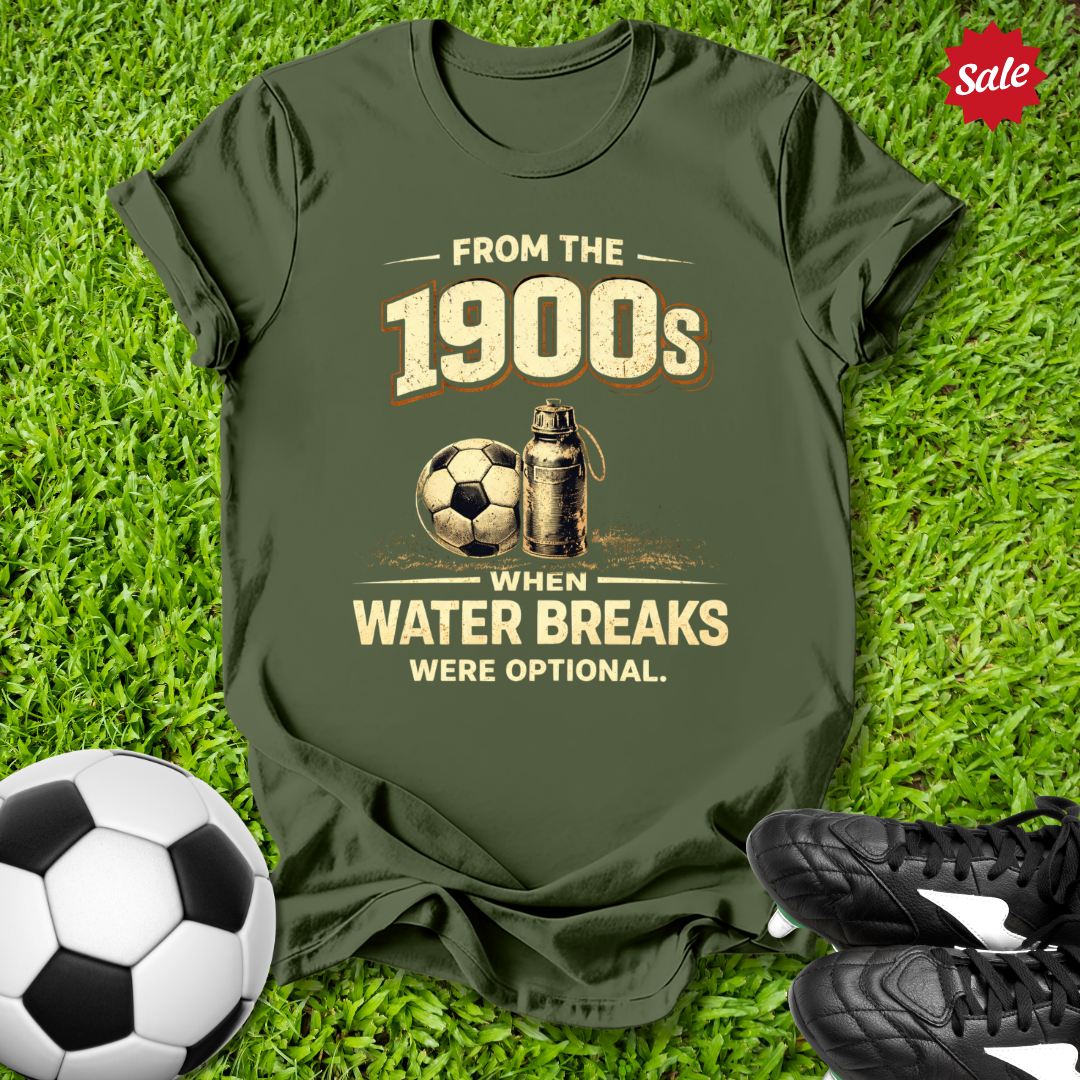 Green t-shirt with soccer-themed design on grass with from the 1900s when water breaks were optional on grass  text  with soccer ball and shoes