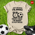 Beige t-shirt with soccer-themed text on grass with soccer ball and shoes