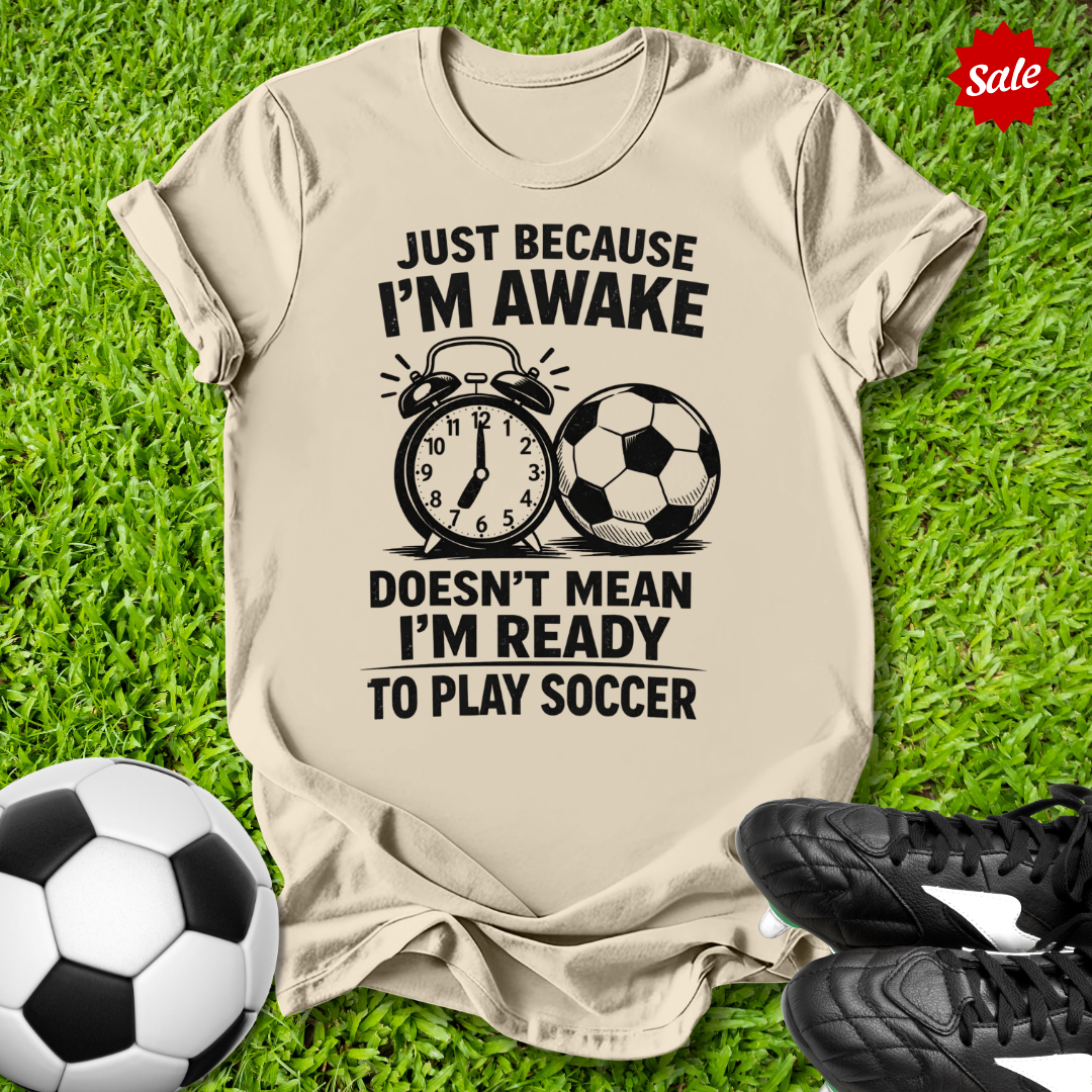 Beige t-shirt with soccer-themed text on grass with soccer ball and shoes
