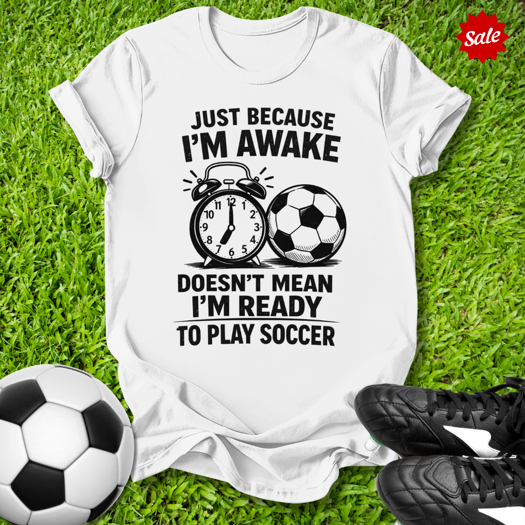 White t-shirt with soccer-themed text on grass with soccer balls and shoes