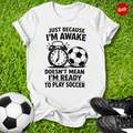 White t-shirt with soccer-themed text on grass with soccer balls and shoes
