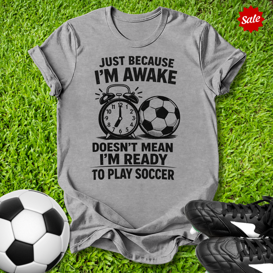 Gray t-shirt with soccer-themed text on grass with soccer balls and shoes